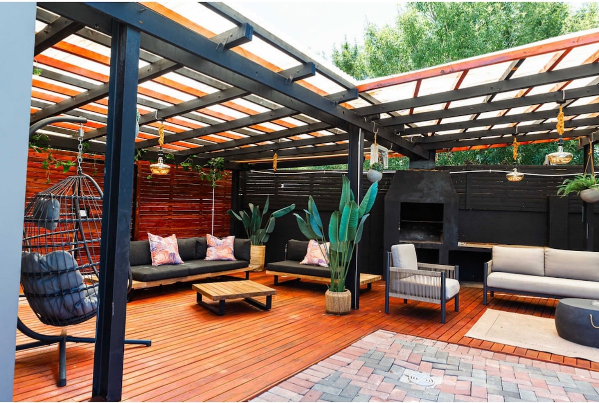An outdoor seating area features a wooden deck surrounded by lush greenery. Comfortable seating options include a hanging chair and multiple couches, complemented by a central wooden coffee table. Decorative lighting fixtures are suspended from the overhead structure, creating a relaxed ambiance.