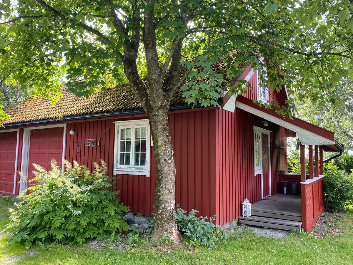 A charming red cottage is framed by lush green foliage, featuring a sloping roof and a covered porch. The exterior is complemented by a white-framed window and a hint of a garage. Grass surrounds the building, creating a serene setting.