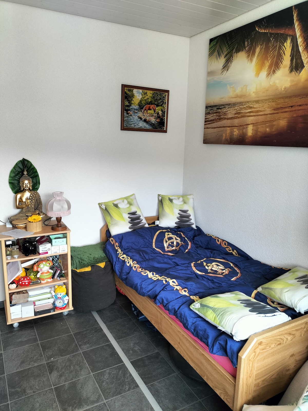 A cozy sleeping area features a wooden bed adorned with patterned pillows and a colorful blanket. A small bookshelf displays various items, while a decorative picture of a tropical scene hangs on the wall. The floor is tiled, contributing to a clean and simple ambiance.
