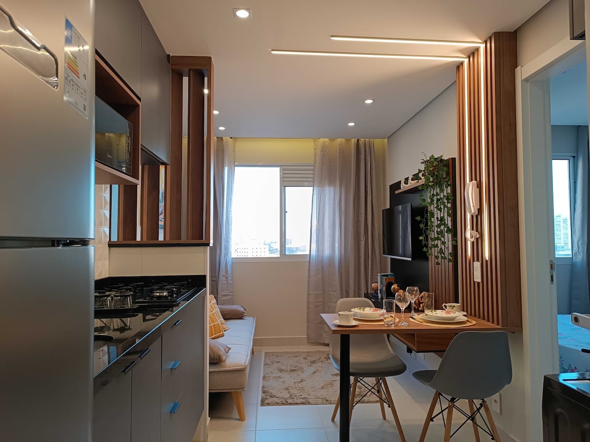 A thoughtfully designed kitchen and dining area features modern cabinetry and sleek appliances. A cozy seating area is positioned beside a large window, allowing natural light to fill the space. A small dining table set for two occupies the center, complemented by tasteful decor elements.