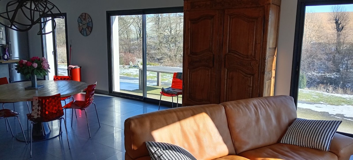 A spacious living area features a comfortable leather sofa, a coffee table, and a contemporary dining table surrounded by vibrant red chairs. Large glass doors allow natural light to fill the room, offering views of the outdoor landscape and terrace.