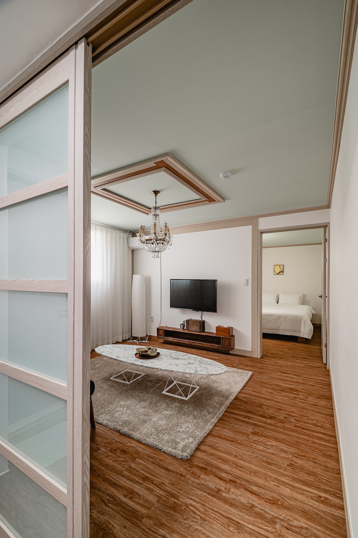 A spacious living area is visible through a glass-paneled door, showcasing a modern TV mounted on a wooden media console. A plush area rug anchors the space, while soft natural light filters through sheer curtains, enhancing the room's warmth.