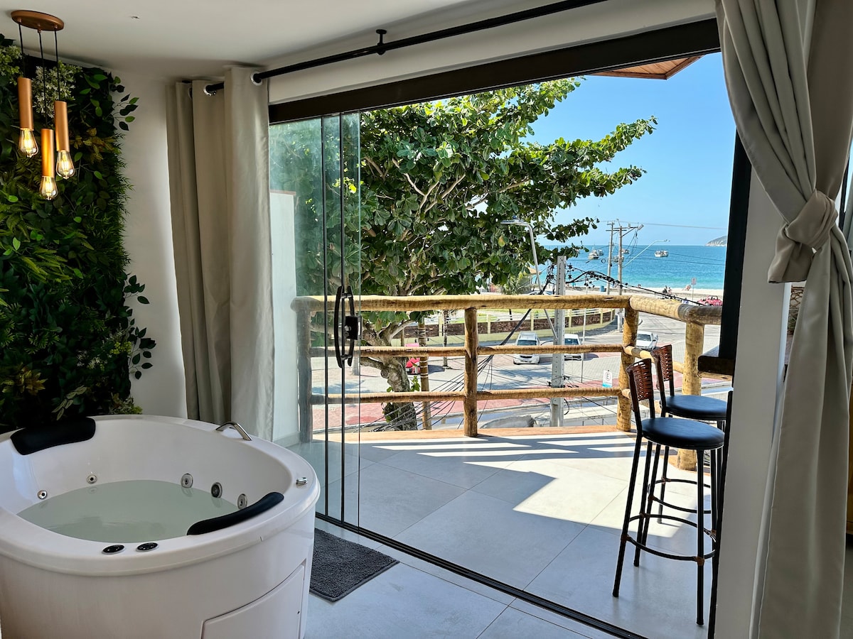 A modern bathroom features a spacious bathtub with a view of the sea through large sliding glass doors. A balcony with wooden railing is visible outside, framed by greenery. Two high chairs are positioned nearby, allowing for relaxation and enjoyment of the coastal scenery.