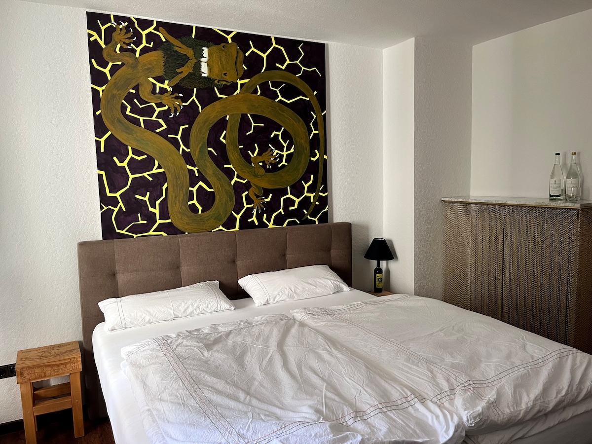 A spacious sleeping area features a large bed with white linens and two pillows. The wall behind the bed is adorned with a vibrant mural depicting a snake against a dark background. A small bedside lamp and a wooden nightstand are placed beside the bed.