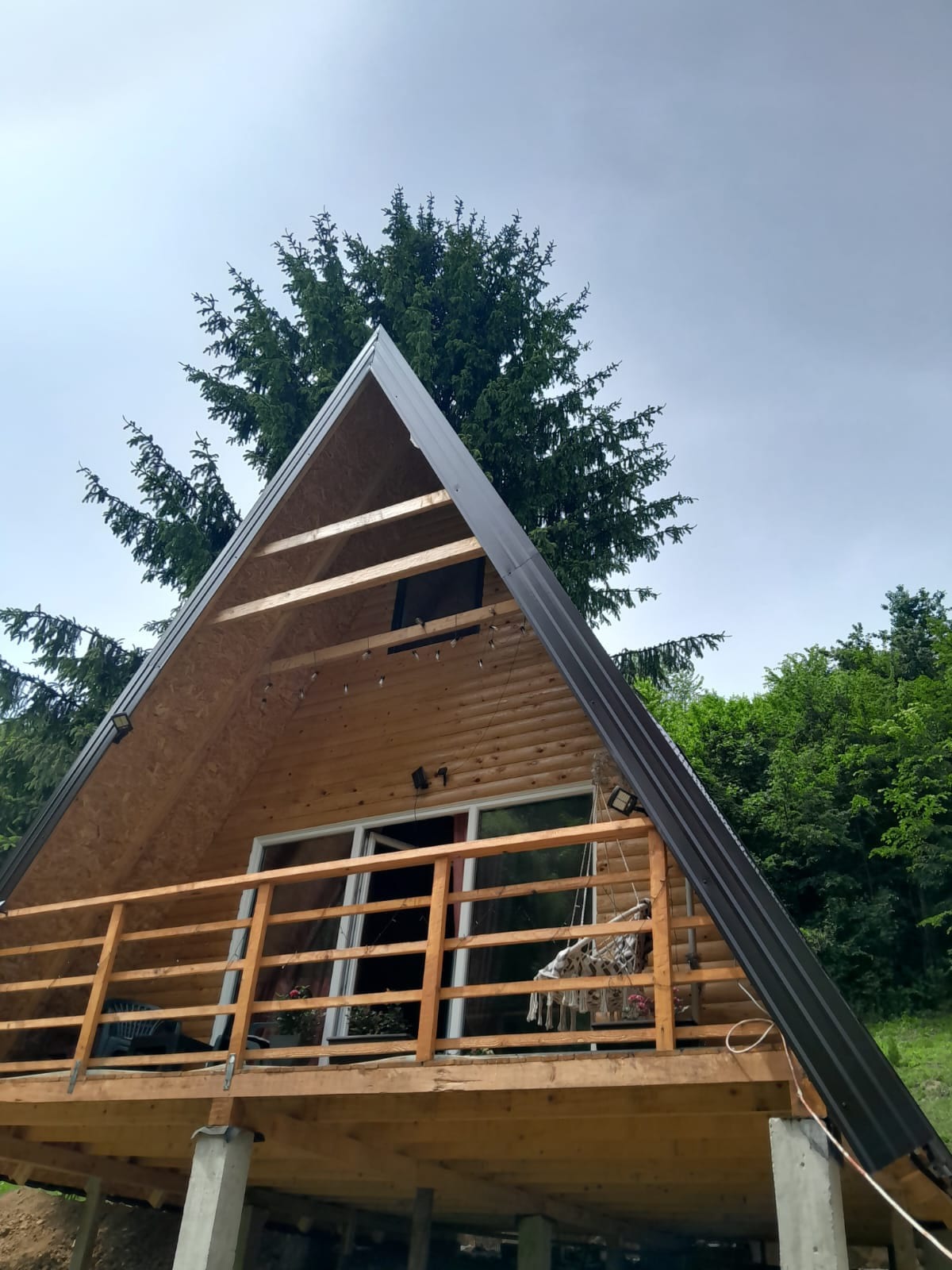 A triangular wooden cabin is showcased, featuring a large deck with a railing. The cabin is surrounded by tall trees, with a few windows visible, allowing natural light to enter. The structure is elevated on sturdy posts, highlighting its unique architecture.