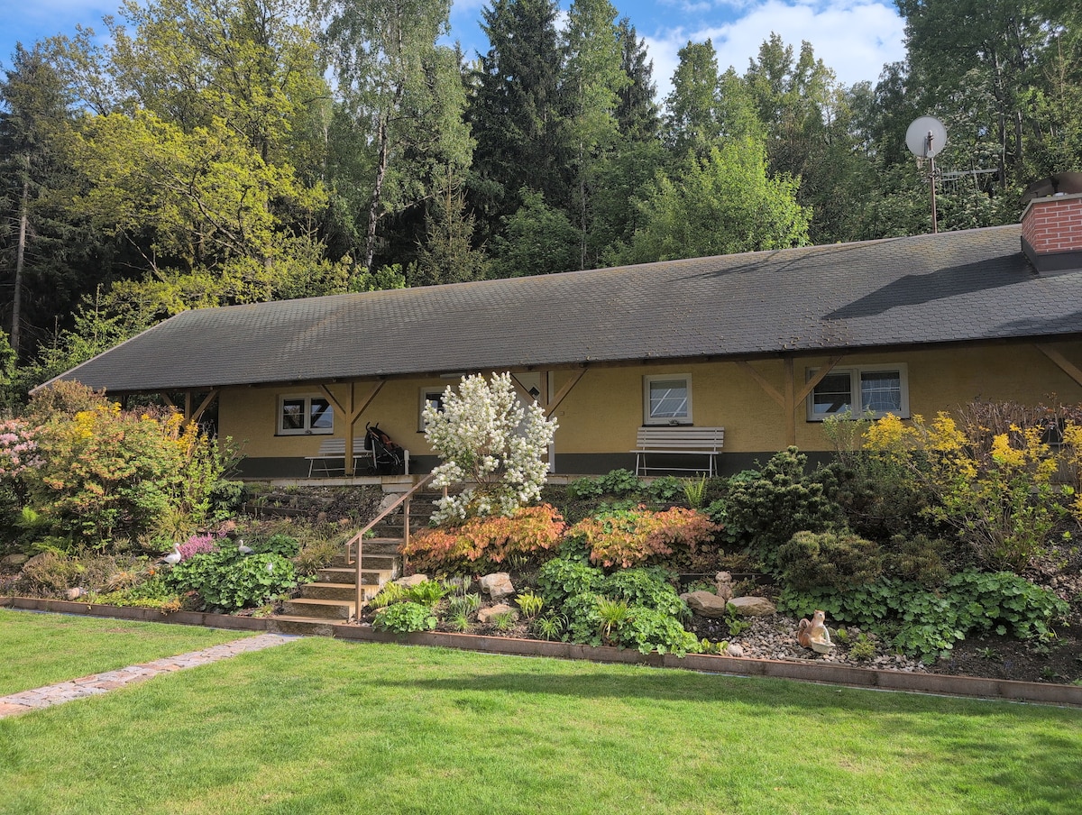 A single-story house is shown surrounded by lush greenery and colorful flowering plants. A set of stone steps leads to the entrance, which is framed by a porch. Large windows enhance the exterior, and a satellite dish is mounted on the roof.