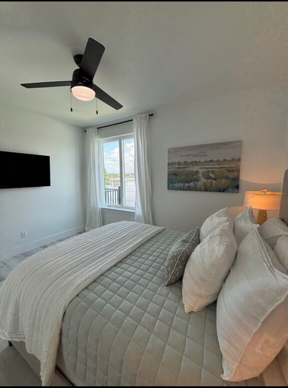 A modern bedroom features a spacious bed adorned with neutral-toned pillows and a light quilt. A soft-lit lamp is positioned on a nightstand beside the bed. Natural light filters through a large window, complemented by sheer curtains, and a wall-mounted television is visible.