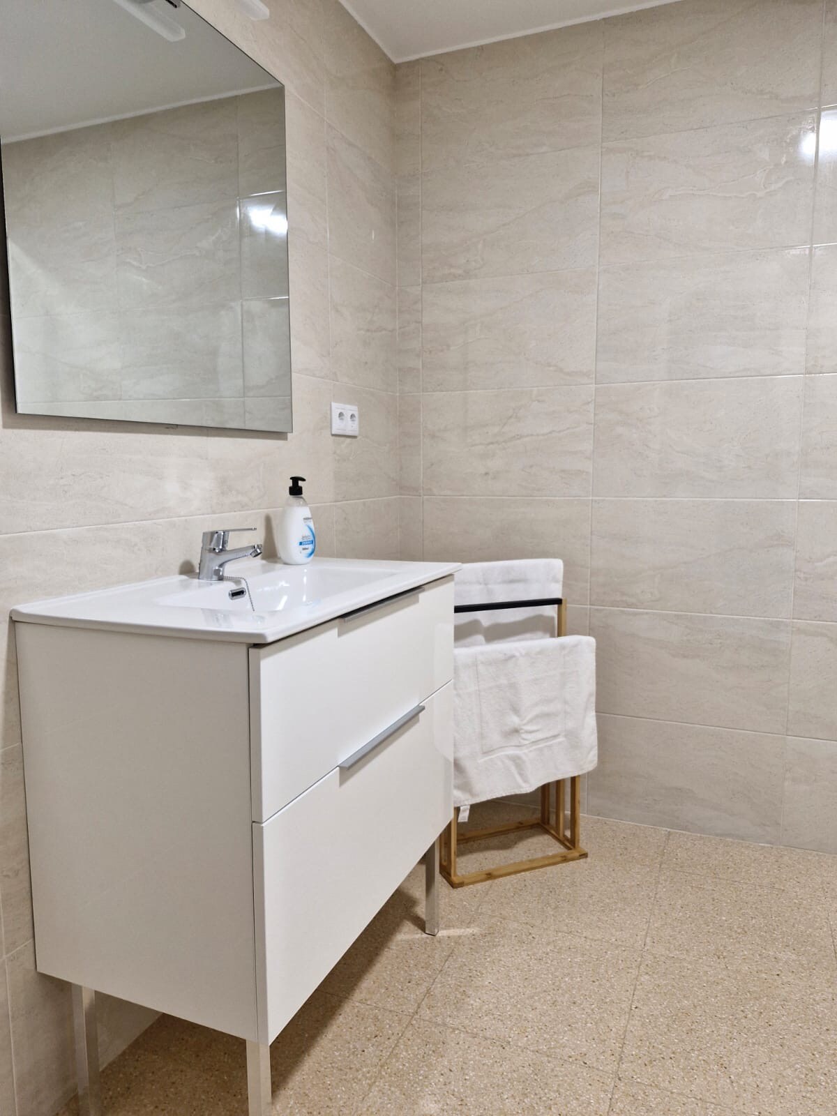 A modern bathroom is presented, featuring a sleek white vanity with a sink and storage drawers. A mirrored cabinet reflects the light, enhancing the bright, tiled space. Two rolled towels are neatly arranged on a golden towel rack, offering convenience and accessibility.