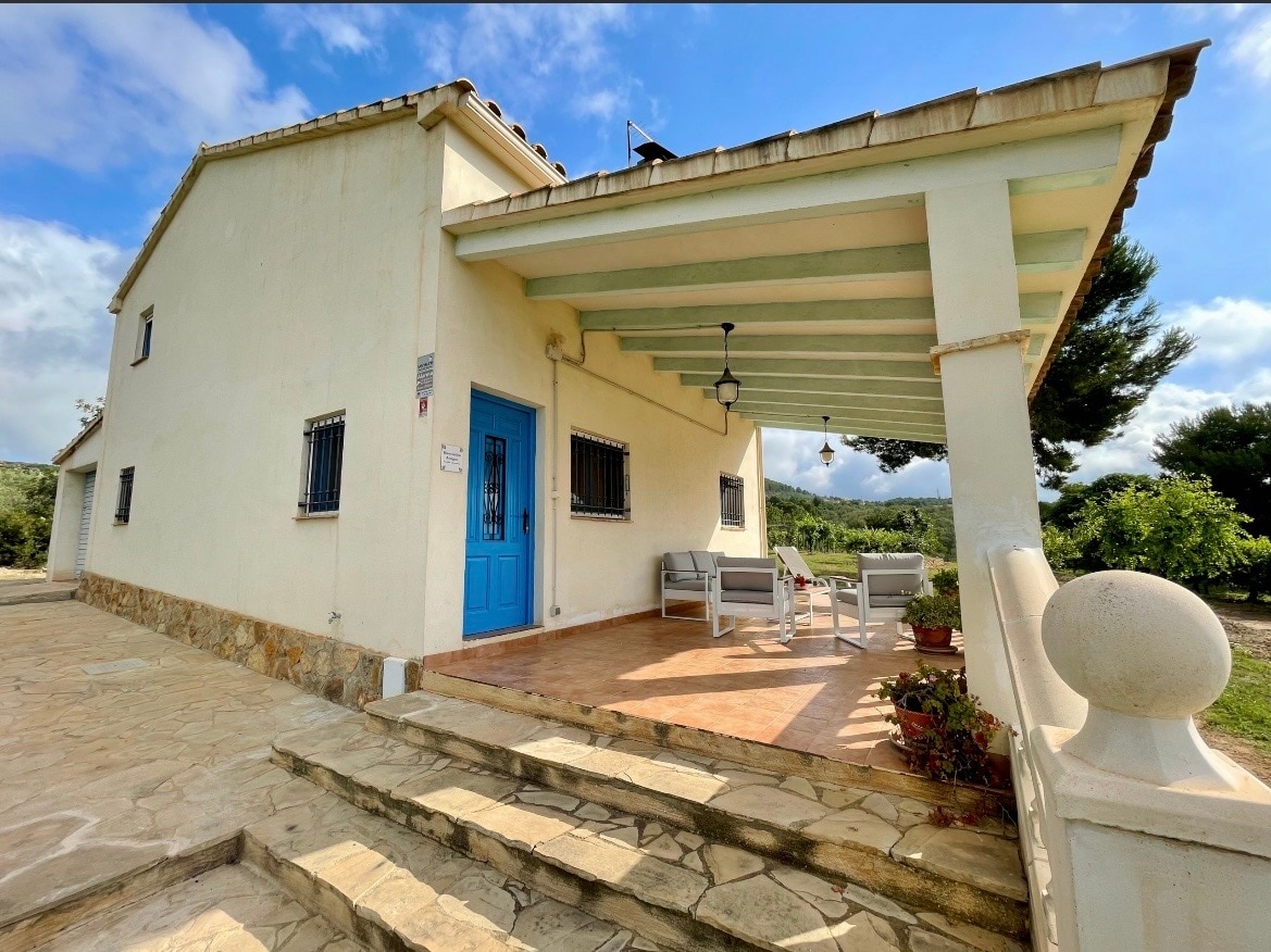 A welcoming porch area is shown, featuring a set of light-colored outdoor furniture arranged on a stone patio. The house has a blue door and is set against a backdrop of trees and greenery, with natural light enhancing the inviting exterior.