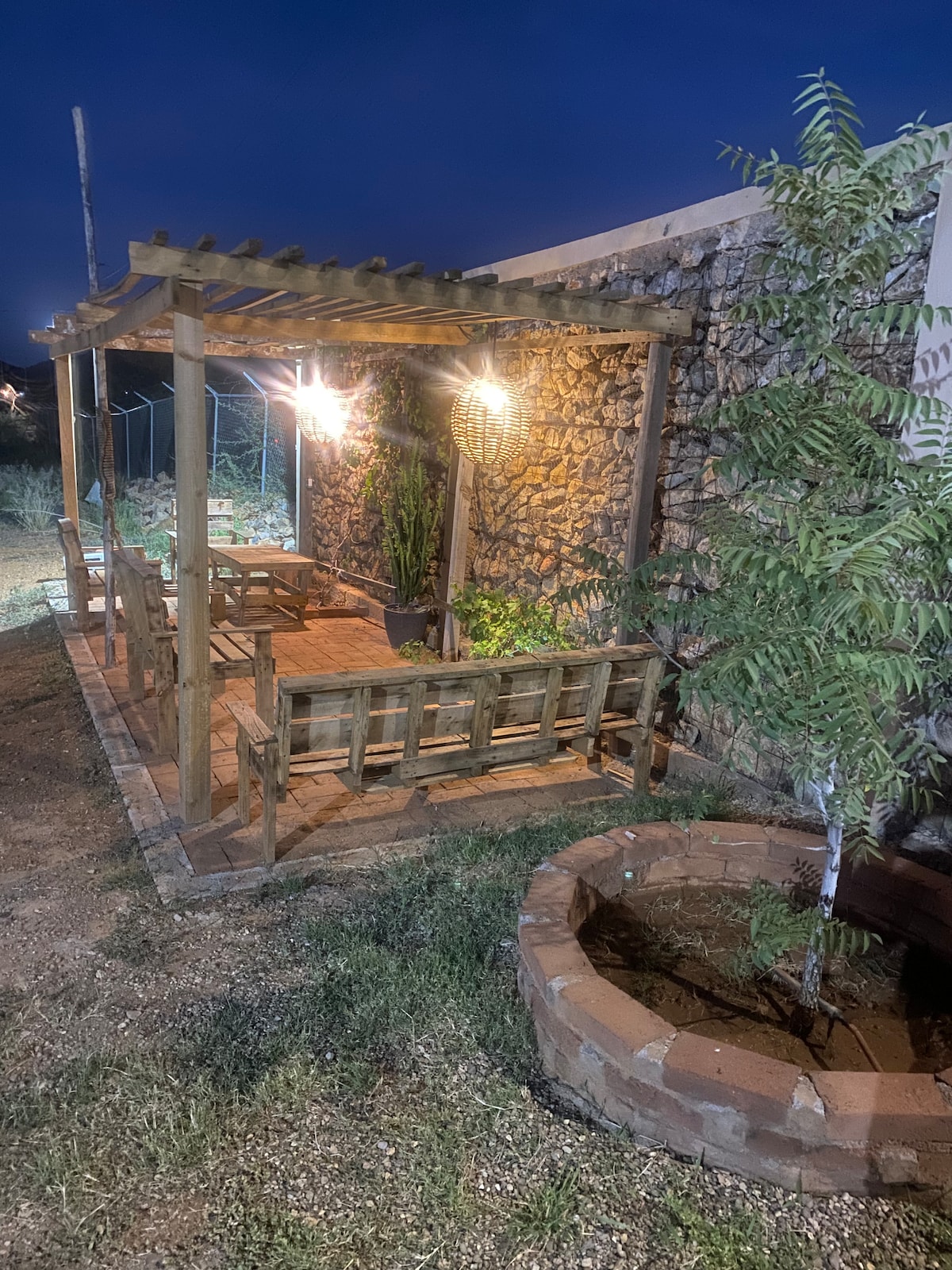 An outdoor seating area is framed by a wooden pergola, illuminated by hanging light fixtures. Stone walls provide texture, while a circular garden bed hosts a small tree. The space is surrounded by a natural landscape, enhancing the connection to nature.