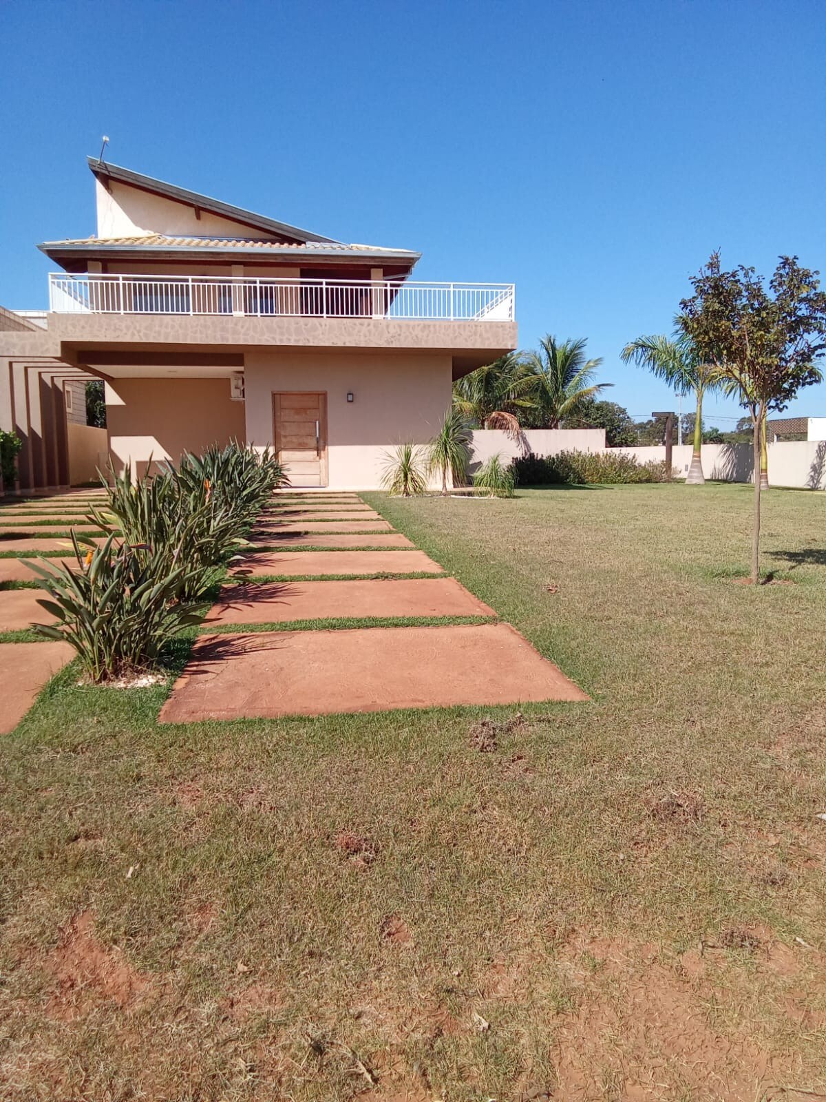 A modern house with a sloped roof is surrounded by a well-maintained lawn. A pathway of wide stone slabs leads to the entrance, framed by symmetrical rows of greenery. Clear skies and palm trees provide a tropical ambiance.