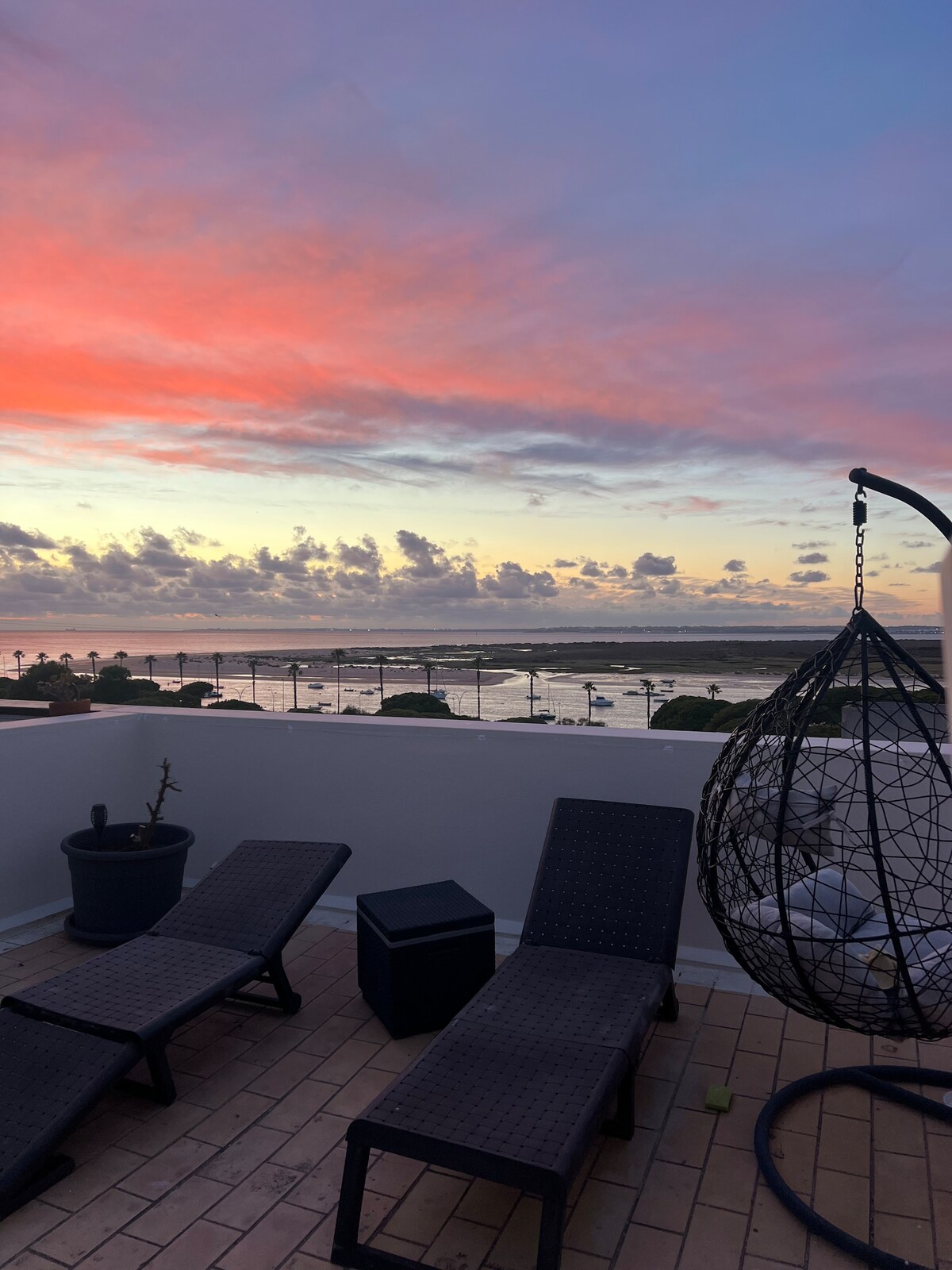 A rooftop terrace is visible, featuring two lounge chairs and a small side table. A hanging chair offers a cozy seating option. The scene is enhanced by a colorful sunset, with soft hues of pink and orange reflected in the sky above the water.