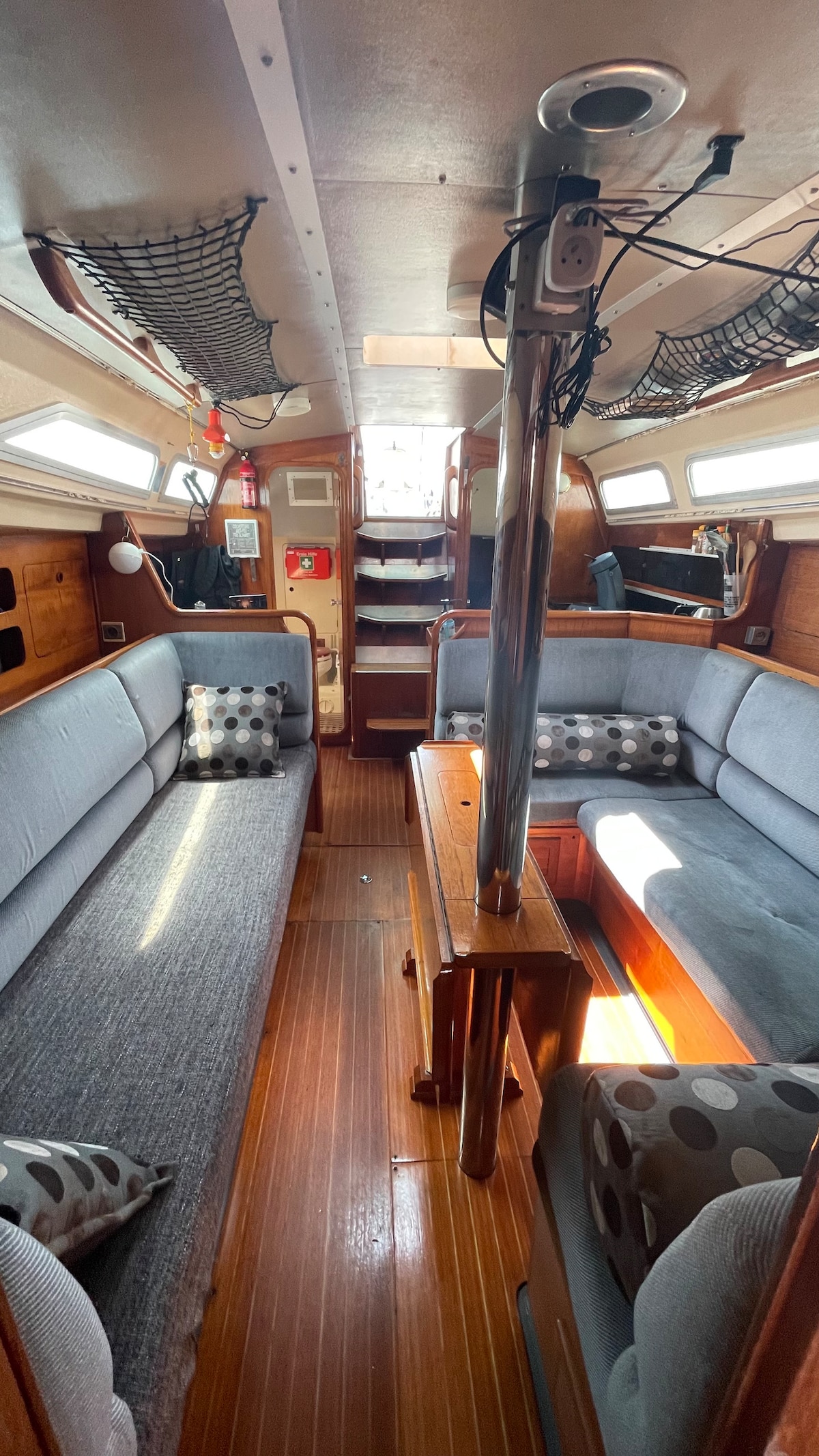 A cozy interior of the sailboat is shown, featuring a central table surrounded by two comfortable gray couches adorned with decorative pillows. Natural light illuminates the wooden accents and creates a warm atmosphere. Storage compartments are visible in the background, along with safety equipment.