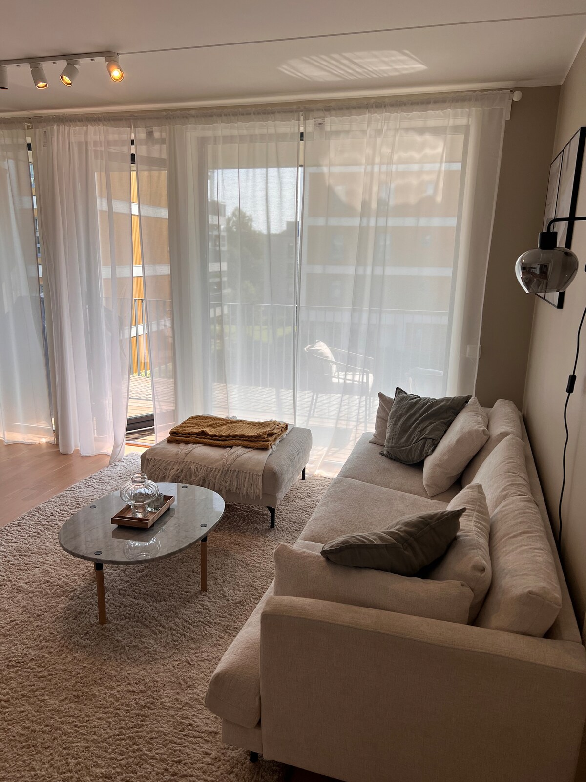A light-filled living area features large sheer curtains allowing natural light to filter through. A cozy beige sofa is positioned facing a glass coffee table, which is topped with a decorative tray. A spacious balcony is visible through the sliding doors.