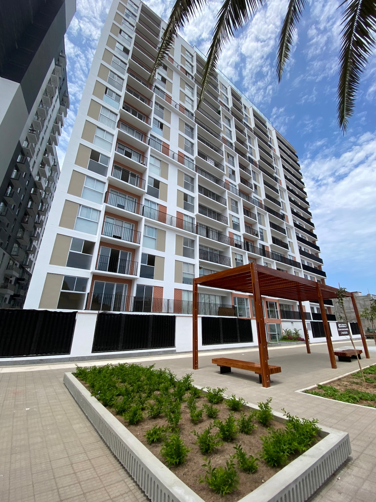 A modern multi-story building is shown, featuring a mix of light and dark exterior tones. Several balconies are visible, framed by large windows. In the foreground, a landscaped area with small shrubs complements a wooden structure designed for seating.