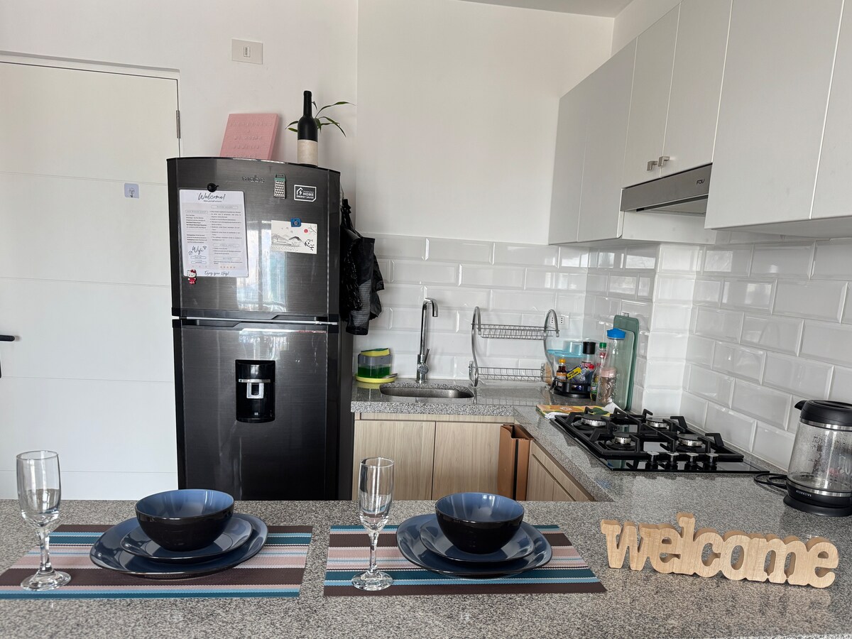The kitchen area features modern appliances and a granite countertop. Two places are set with blue dishes, glasses, and striped placemats. A welcome sign is displayed on the countertop, adding a personal touch to the space.