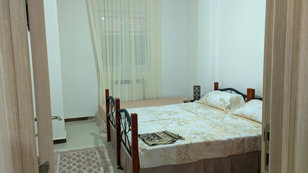 A well-lit bedroom features a spacious bed with a floral-patterned coverlet. Soft curtains allow natural light to filter in, highlighting the neutral tones of the walls and furnishings. A decorative area rug adds comfort to the floor, contributing to a serene environment.