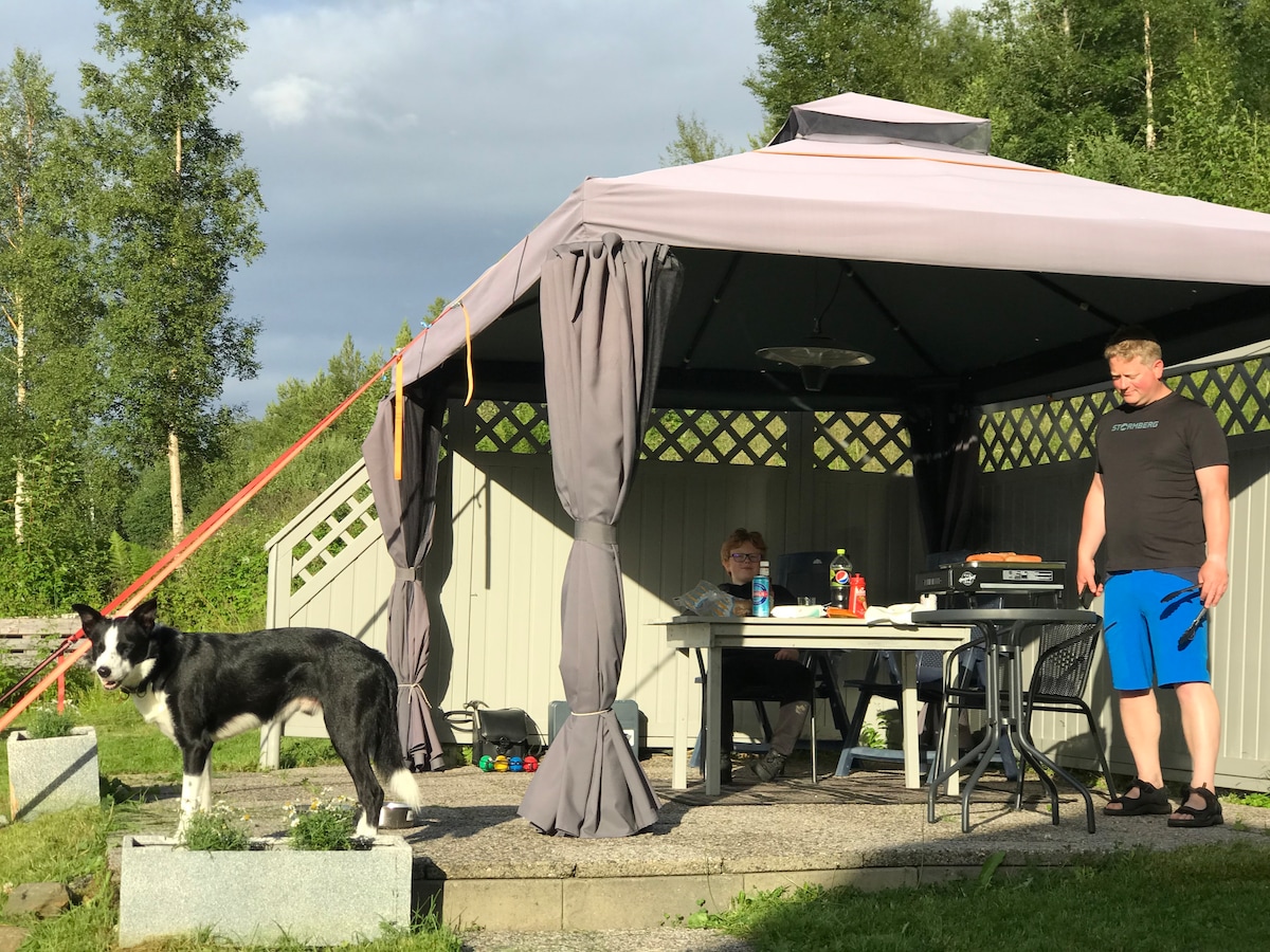 A shaded pavilion with a light-colored roof is set among greenery. A table and chairs are arranged underneath, while a black and white dog stands on a stone base nearby. Two individuals are seen engaged in conversation, surrounded by an atmosphere of relaxation.