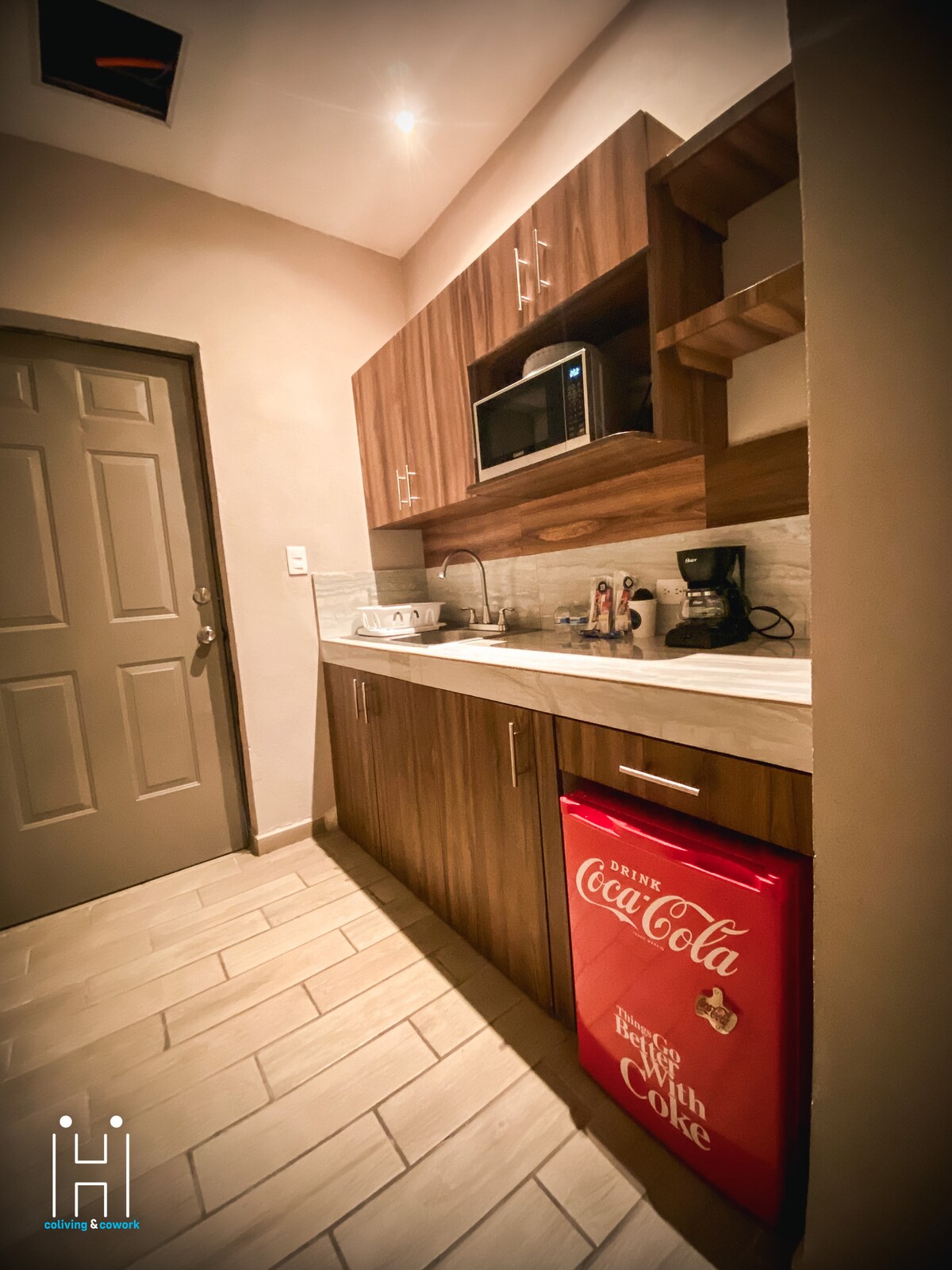 A compact kitchen area is presented, featuring wooden cabinets and a countertop. A microwave and coffee maker are placed prominently. A red Coca-Cola mini fridge adds a pop of color, while the entrance door and light-colored walls enhance the overall illumination.