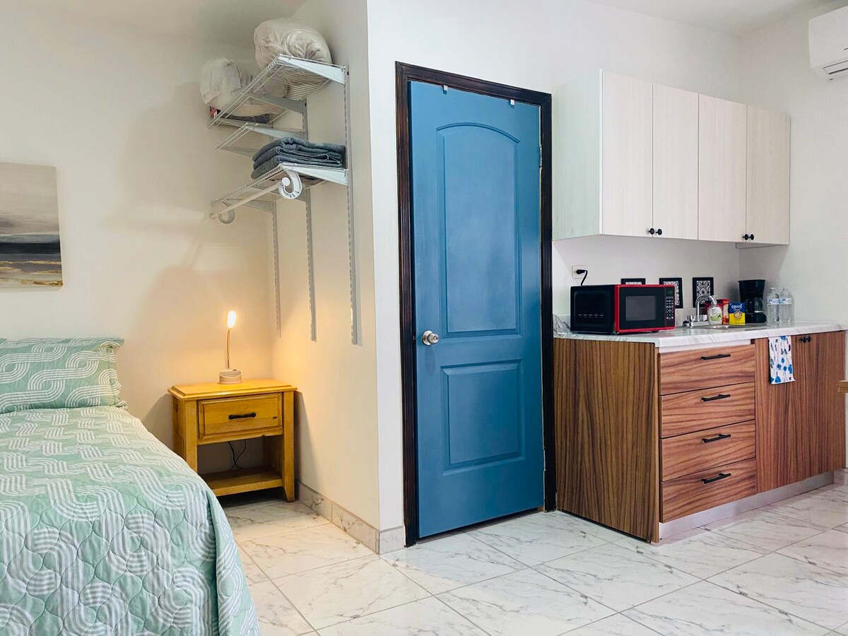 A simple bedroom area features a bed with a light green coverlet, complemented by a nightstand with a lamp. A blue door provides access to the room, while a kitchenette with wooden cabinetry is seen in the corner, equipped with a microwave and coffee maker.