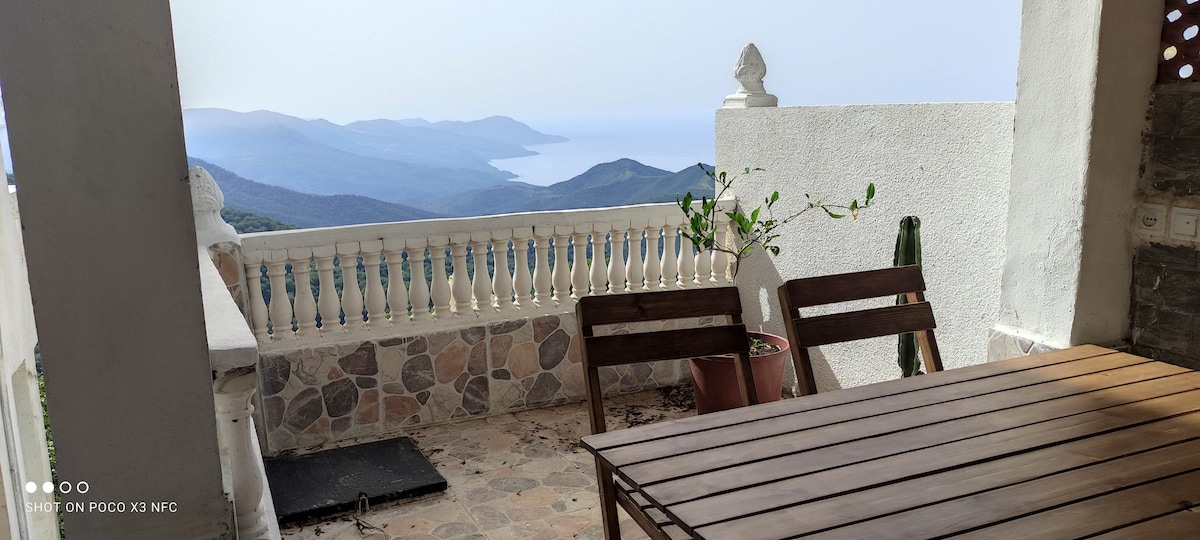 A spacious balcony offers a stunning view of rolling hills and a distant body of water. Wooden furniture is arranged neatly, while potted plants add a touch of greenery. The balustrade is crafted from stone, enhancing the outdoor space's ambiance.