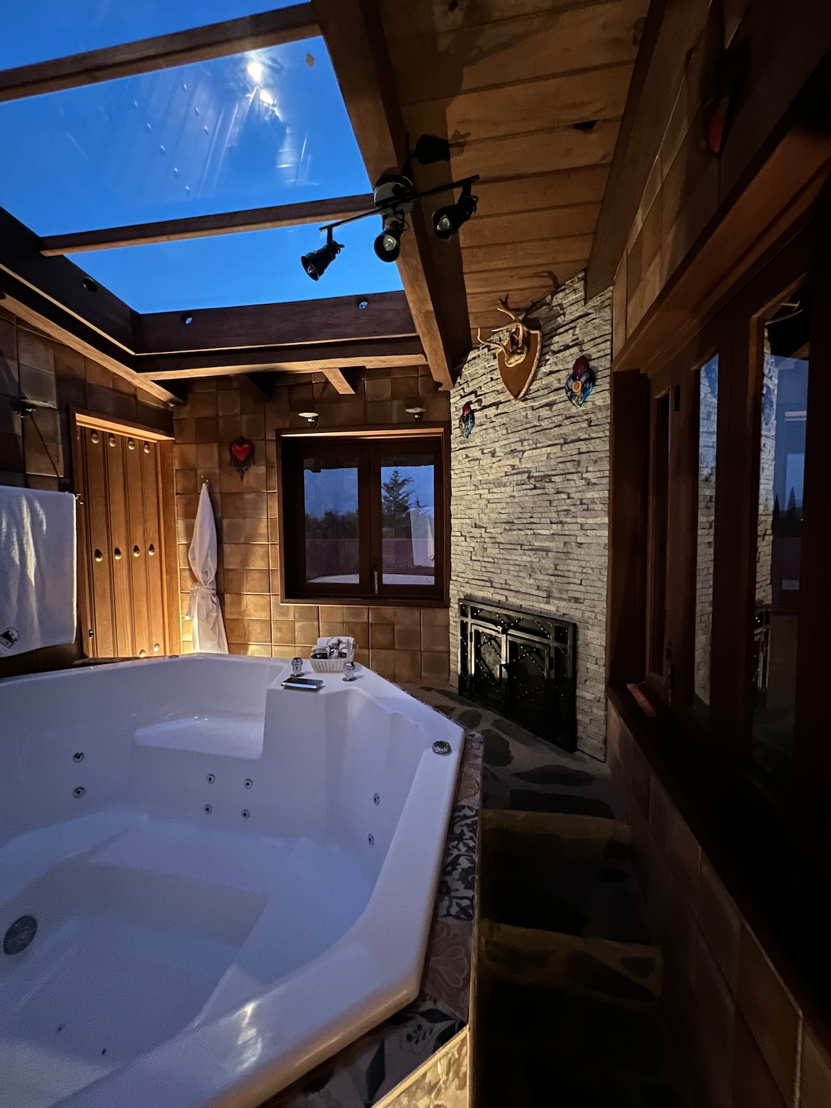 A spacious jacuzzi is located in a warmly lit bathroom featuring wooden beams and stone accents. A large window offers nighttime views of the sky, while soft lighting enhances the natural materials. Towels are neatly arranged nearby, contributing to a serene atmosphere.