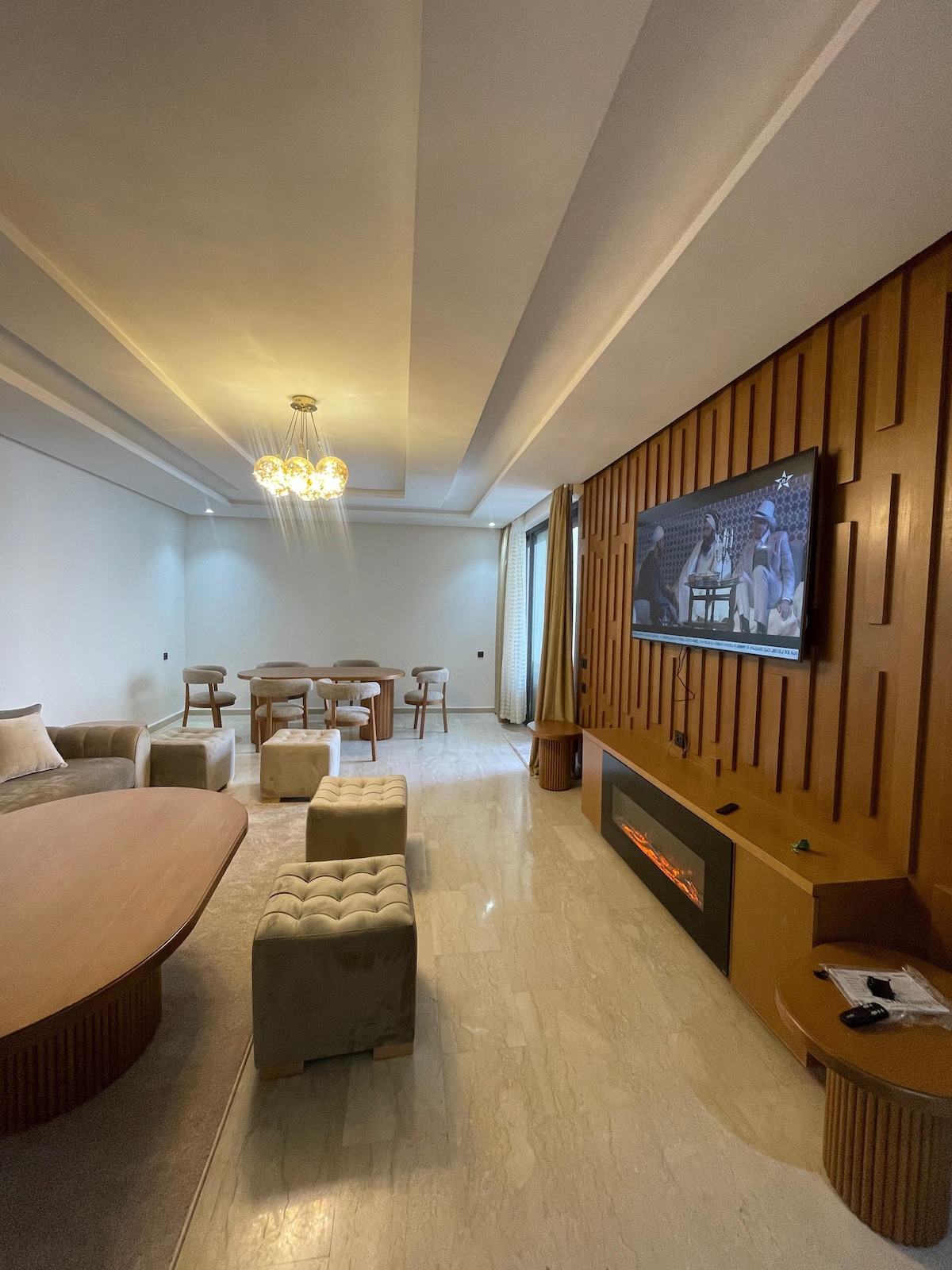 A spacious living area is highlighted by a large oval table surrounded by soft, cushioned stools and accent chairs. A modern entertainment unit features a flat-screen TV, while a warm glow emanates from an electric fireplace beneath it. Natural light enters through the window, enhancing the neutral decor.