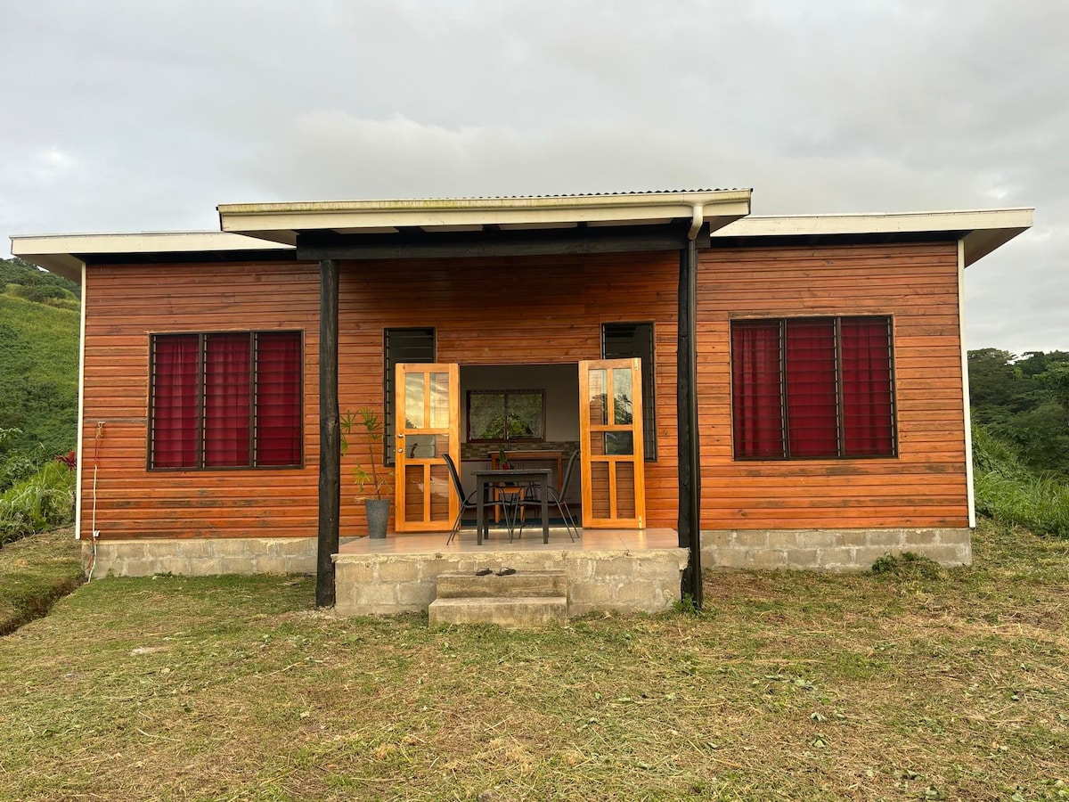 A wooden exterior of a house features large double doors, which are open to reveal a welcoming entry. Two windows with red curtains complement the design. A small flight of steps leads to the entrance, and greenery is visible in the background.