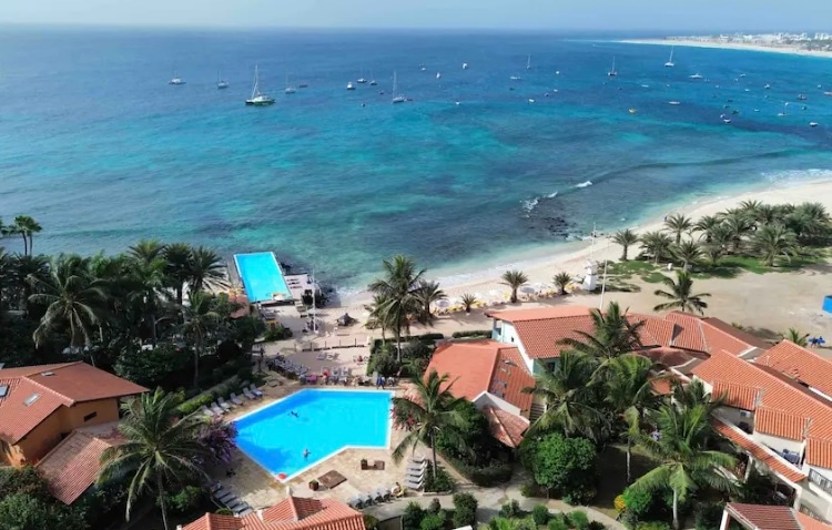 An aerial view captures the serene coastline, with a vibrant blue pool surrounded by palm trees. The beach and clear waters are highlighted, while boats are seen floating gently in the distance. Brightly colored rooftops of nearby buildings complement the coastal scenery.