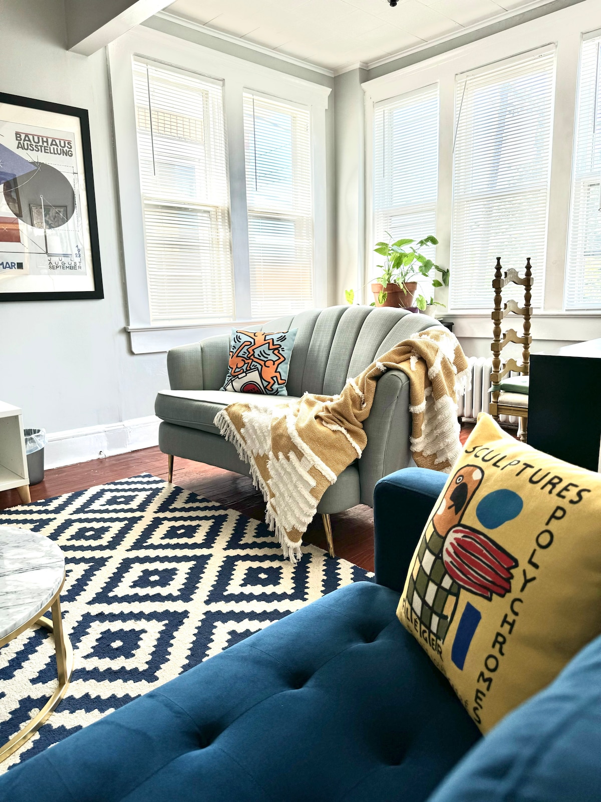 A bright living room features a plush gray sofa adorned with a colorful decorative pillow and a soft throw draped casually. A patterned area rug anchors the space, complemented by a sunny corner filled with greenery and large windows that allow natural light to fill the room.