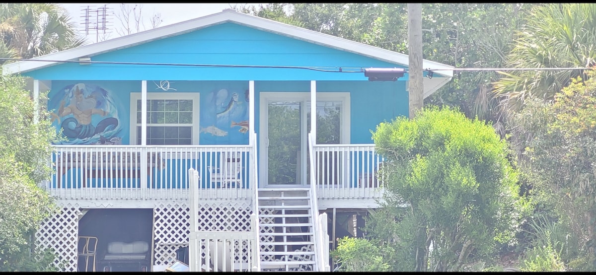 A charming bayfront house with a vibrant blue exterior is depicted. A spacious deck with white railings is accessed by a set of outdoor stairs. Tropical greenery surrounds the home, while colorful murals are painted on the walls, adding character to the inviting entrance.