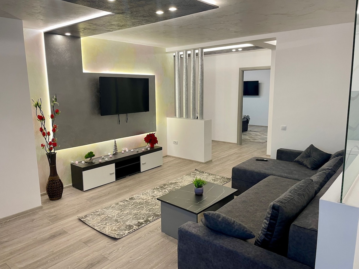 A comfortable living area is present, featuring a dark gray sofa positioned around a low coffee table. A flat-screen TV is mounted on a softly illuminated accent wall. Decorative elements include a potted plant and artificial flowers, enhancing the modern aesthetic of the space.