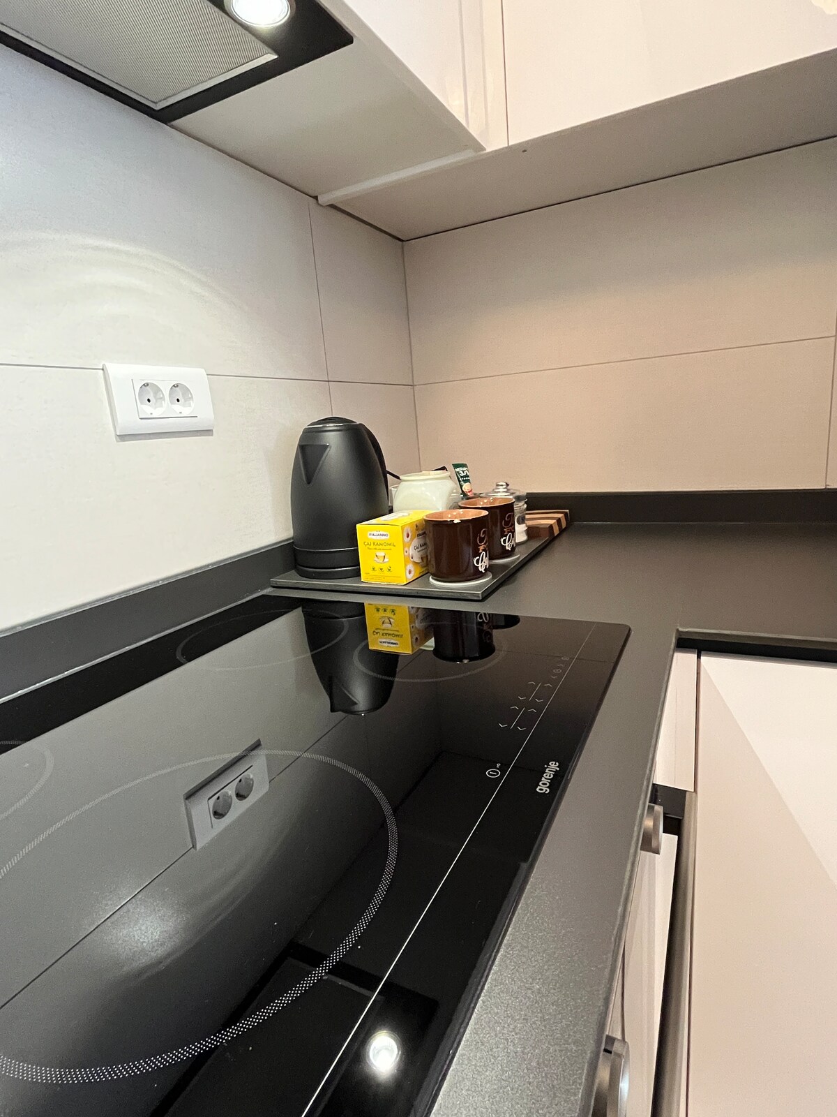 The modern kitchen features a sleek countertop with an induction cooktop. An electric kettle and a tray of various condiments, including coffee and tea, are arranged neatly. The cabinetry is minimalist, enhancing the clean lines of the space.