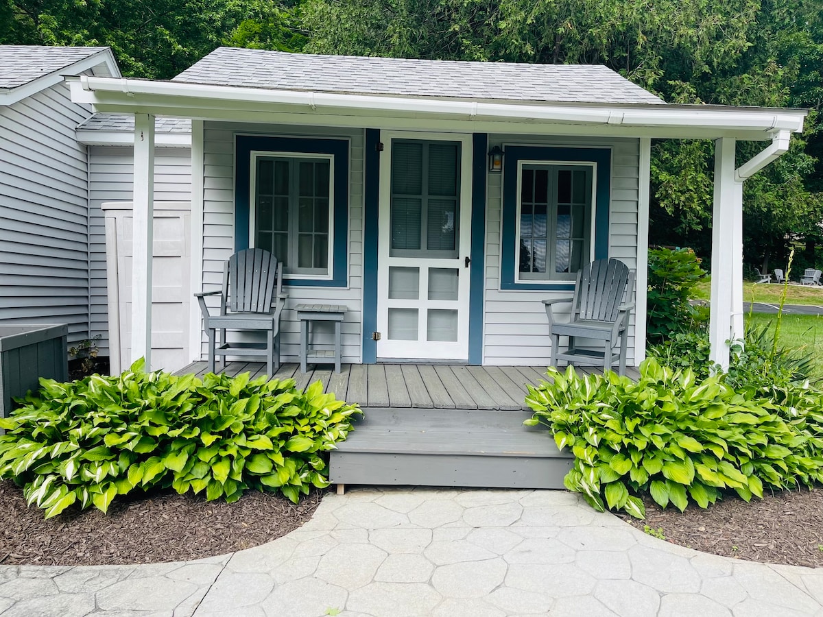 A charming cottage entrance is shown, featuring a covered porch with two wooden chairs set against a backdrop of well-maintained greenery. The door is framed by large windows, inviting natural light into the space. A pathway made of stone leads up to the cottage.