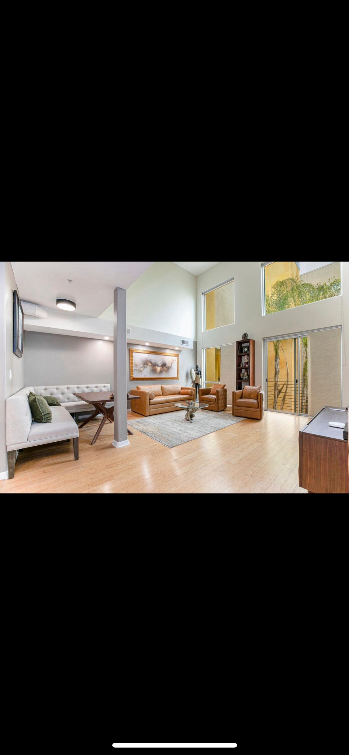 A bright living area is presented with high ceilings and large windows allowing natural light. The space features a stylish dining table and chairs alongside a comfortable leather sofa. Tall windows reveal exterior greenery, enhancing the relaxed ambiance.
