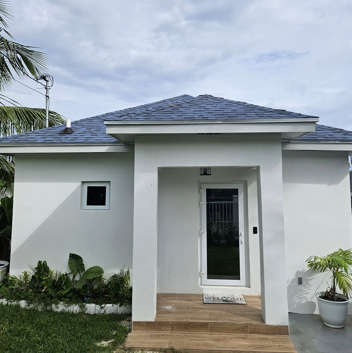 The exterior of a charming cottage features a clean white facade and a blue-shingled roof. A welcoming front door is framed by a tiled entryway, while small plants are positioned along the sides, contributing to an inviting appearance.