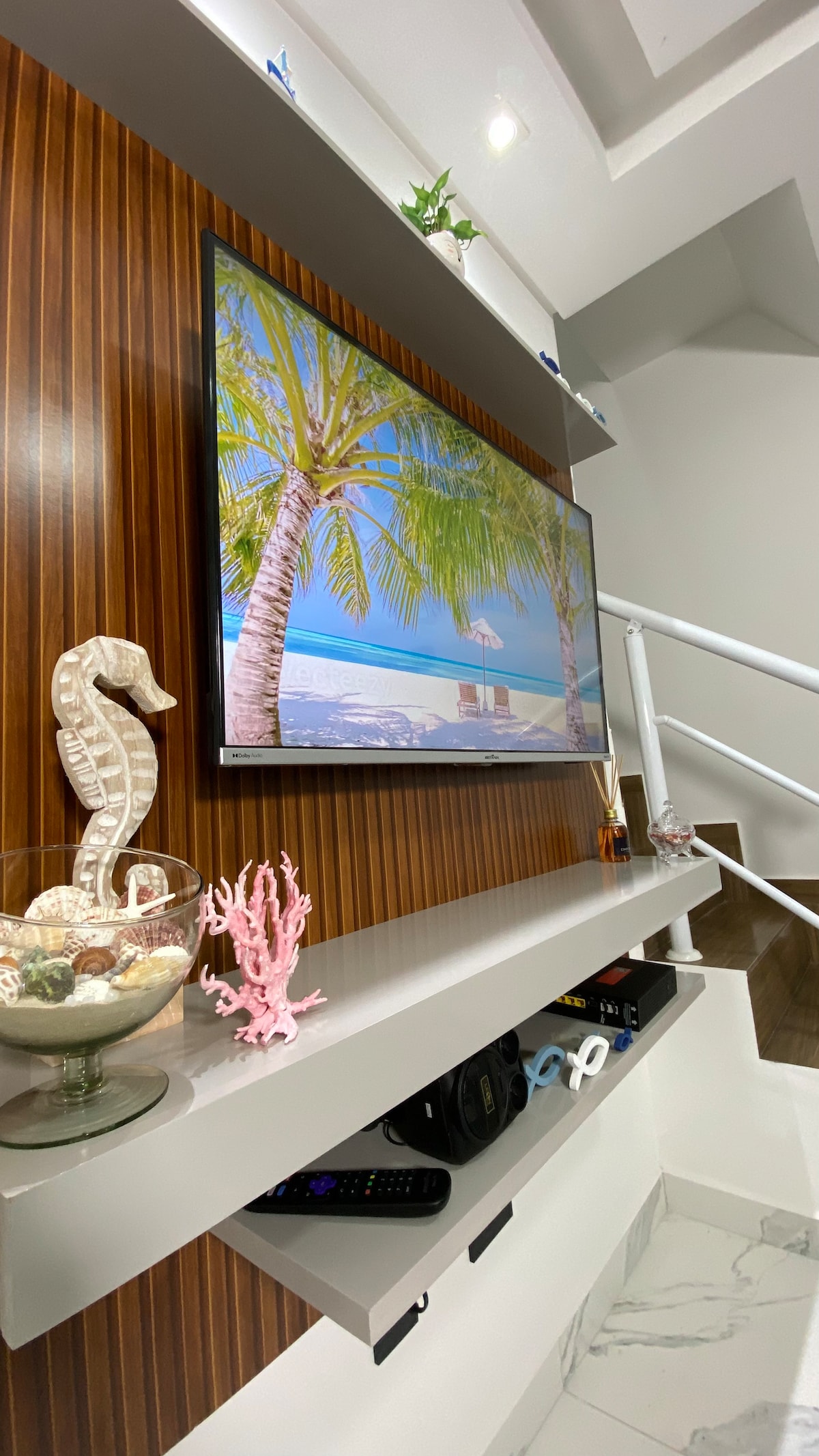 A sleek entertainment area is highlighted by a mounted TV displaying a serene beach scene. Below, a decorative shelf holds a glass bowl with seashells and coral accents. The backdrop features warm wooden paneling, adding a touch of elegance to the space.