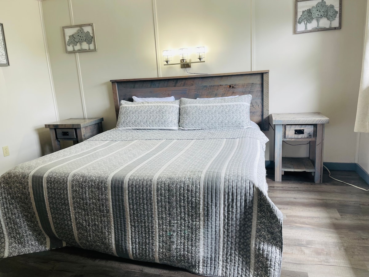 A spacious sleeping area is presented, featuring a king bed with a striped quilt and textured pillows. Flanking the bed are two nightstands equipped with lamps. Soft lighting contributes to a calm ambiance, while light filters through nearby window coverings.