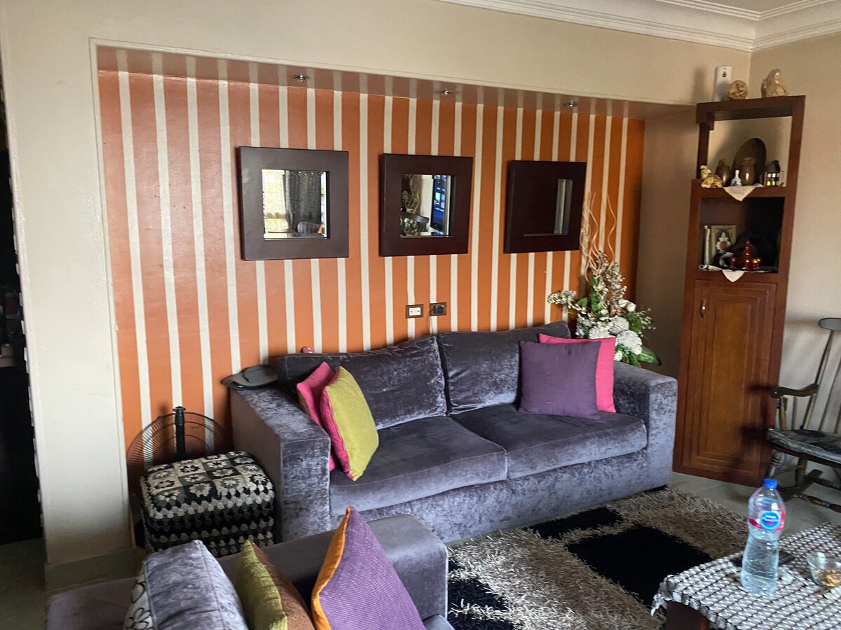 A modern living area features a plush gray sofa adorned with vibrant purple and yellow cushions. The wall is decorated with vertical orange stripes and three mirrors, while a wooden shelf displays decorative items. A small glass table complements the space.