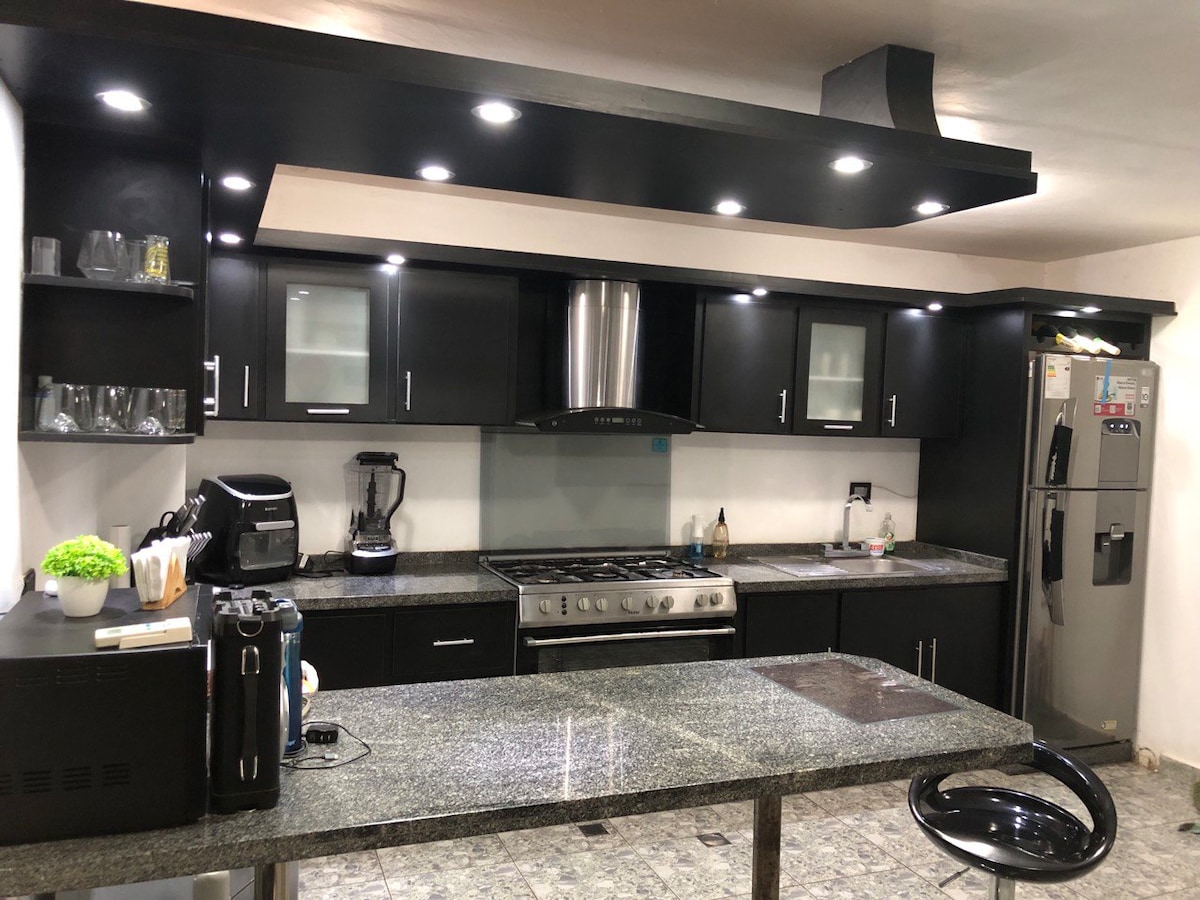 A modern kitchen is presented with sleek black cabinetry and a spacious granite countertop. Under-cabinet lighting illuminates the area, enhancing visibility. Stainless steel appliances are prominently featured, with a refrigerator and a coffee machine among the amenities. The flooring is composed of light-colored tiles.