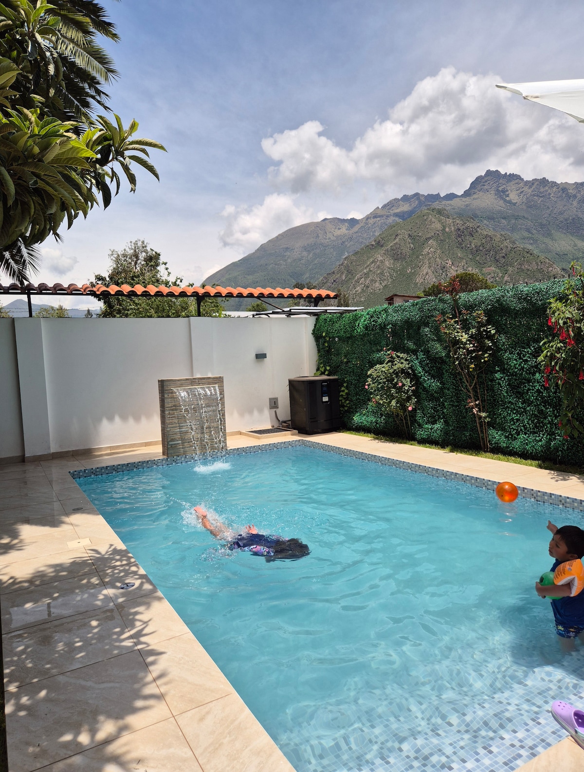 The image depicts a tranquil swimming pool surrounded by lush greenery, bordered by a white wall. A cascading waterfall feature adds a soothing element. The scenic backdrop includes mountains under a clear sky, enhancing the outdoor relaxation experience.