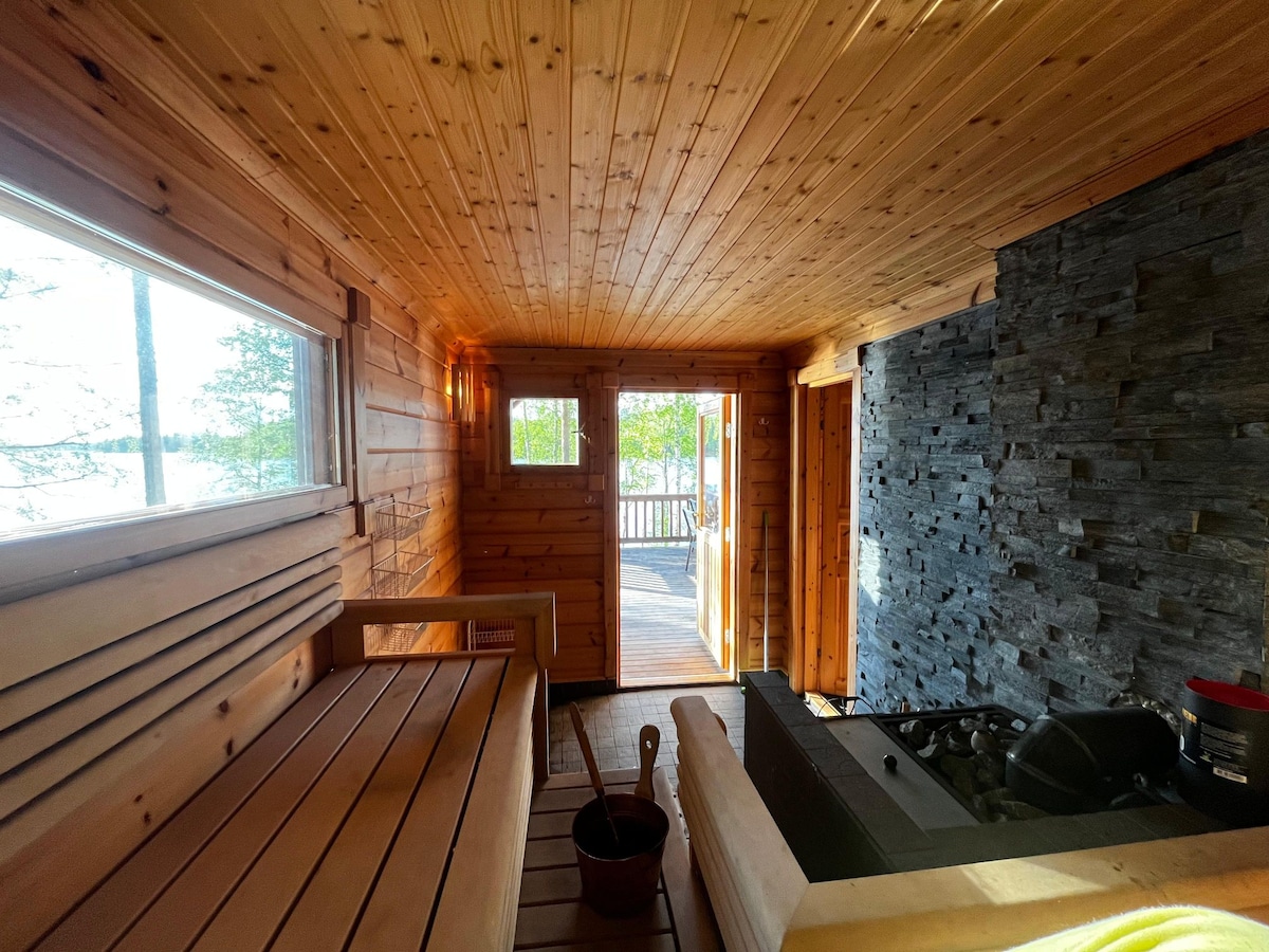 The interior of the sauna is lined with warm wood, featuring a built-in bench. A window allows natural light to illuminate the space, while the entrance leads to an outdoor deck. A stone wall adds texture, enhancing the rustic charm of the setting.