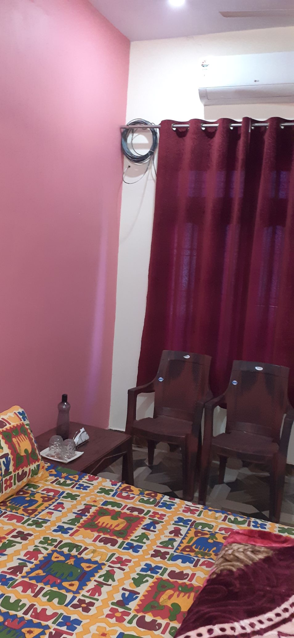 A cozy room features a vibrant pink wall, complemented by maroon curtains that frame a window. Two wooden chairs are positioned beside a small table, which holds a bottle and a decorative item. A colorful patterned throw adorns the bed.