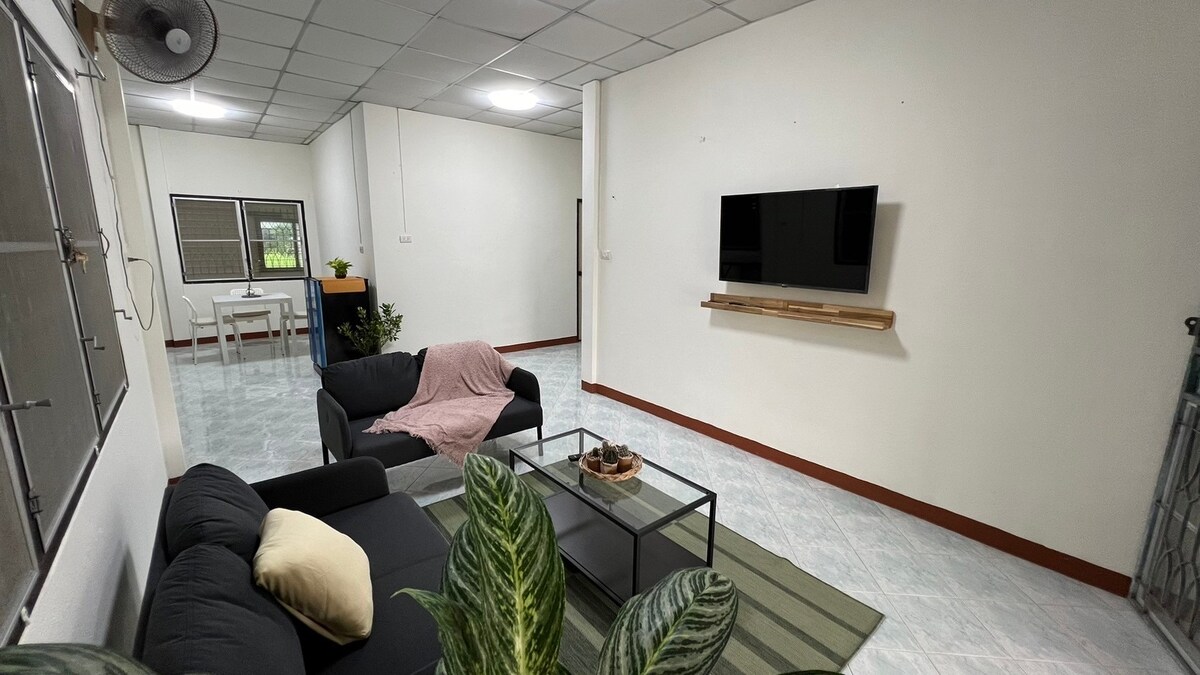 A spacious living area showcases a black sofa set, accented by a light throw blanket and a decorative pillow. A glass coffee table sits in the center, while a wall-mounted television provides entertainment. Natural light enters through the windows, enhancing the room's openness.
