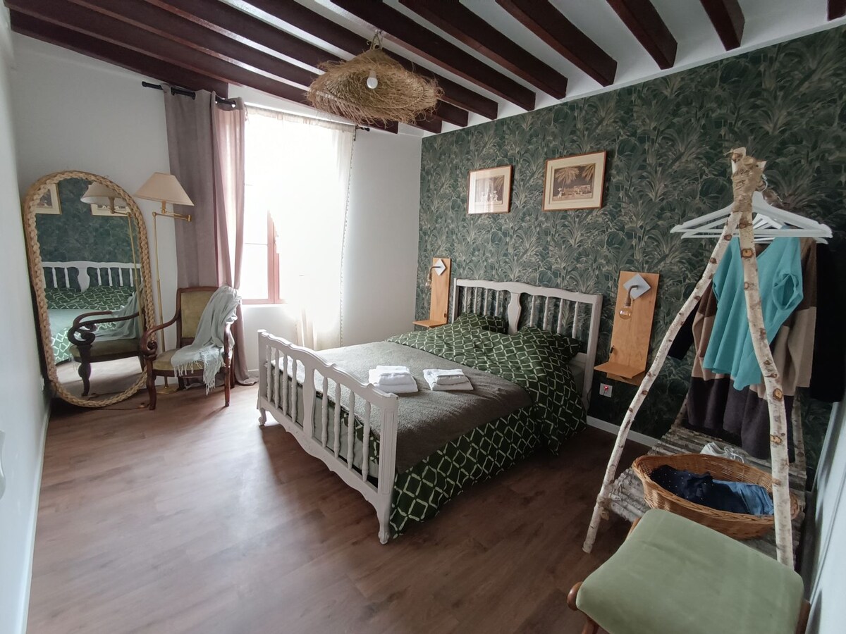 A serene bedroom is equipped with a spacious double bed, adorned with patterned bedding. A mirror and a soft armchair accent the room, complemented by a wooden clothing rack displaying garments. Natural light filters in through the window, highlighting the botanical wallpaper and exposed beams.