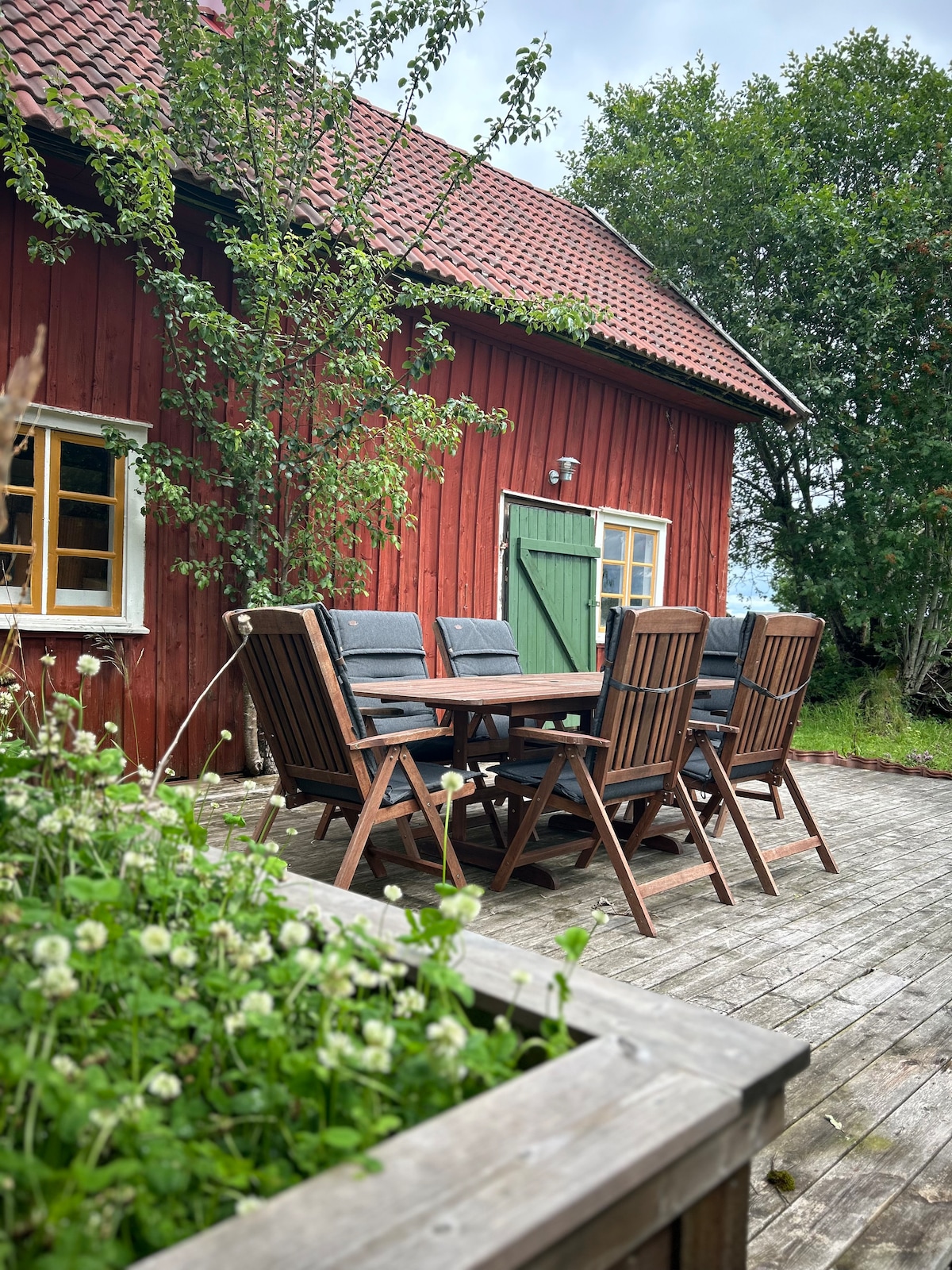 A spacious outdoor dining area features a wooden table accompanied by six comfortable chairs. The red wooden house serves as a backdrop, with green grass and blooming plants surrounding the space. A tree nearby adds natural shade to the area.
