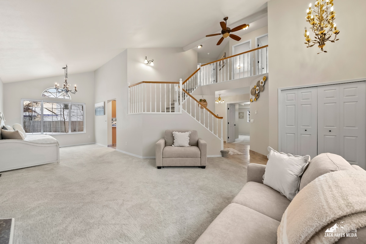 The spacious living area features light-colored carpeting and neutral walls. A large window allows natural light to fill the space. A decorative staircase leads to an upper level, while a cozy sofa and stylish light fixtures add to the overall ambiance.