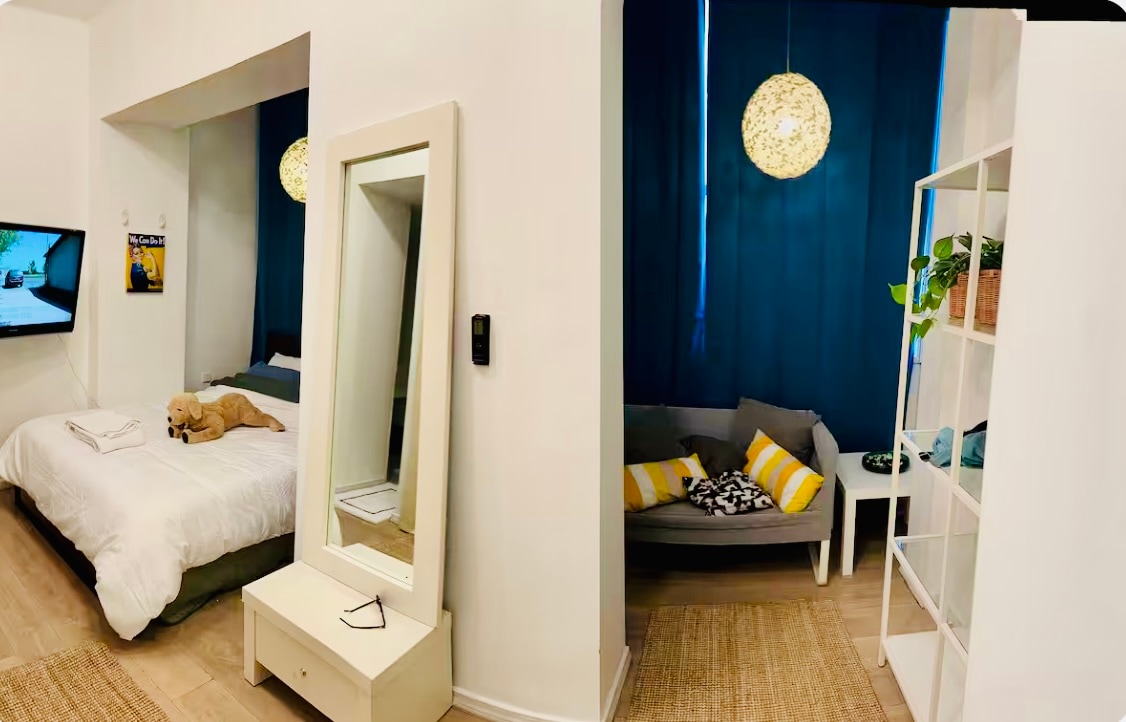 A bright sleeping area is showcased with a spacious bed, accompanied by a large mirror and a stylish floor lamp. A cozy seating area is visible, featuring a gray sofa adorned with decorative cushions. Soft blue curtains create a soothing atmosphere.