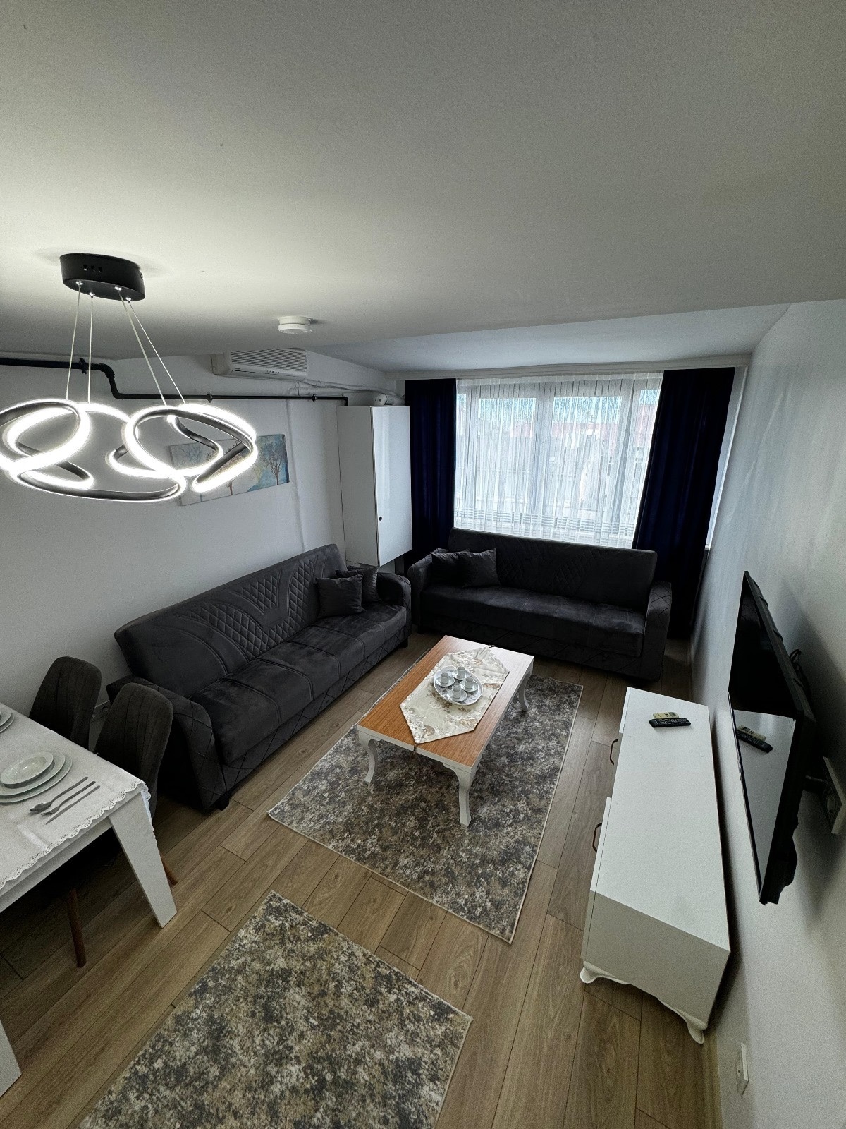 A modern living area is presented, featuring two gray sofas positioned around a central coffee table. A stylish light fixture hangs above. A television is mounted on the wall opposite the seating area, and large windows adorned with dark curtains allow natural light to fill the space.