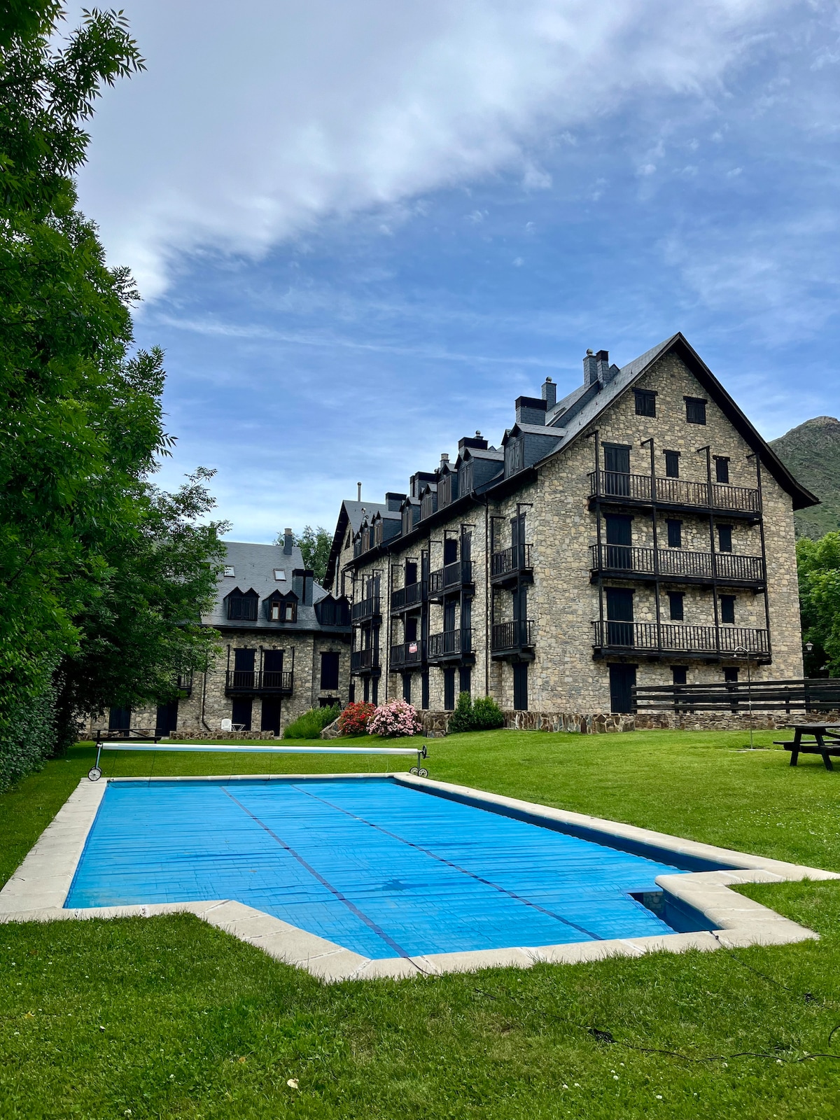 An outdoor view of the accommodation is presented, showcasing an expansive lawn with vibrant greenery and a freshly covered pool. The multi-story stone building features multiple balconies and dark roofing, surrounded by trees and landscaped areas, indicating a serene and spacious environment.